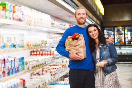 笑脸夫妇与纸杂货袋充满产品快乐地看着相机在现代超市乳制品部门照片